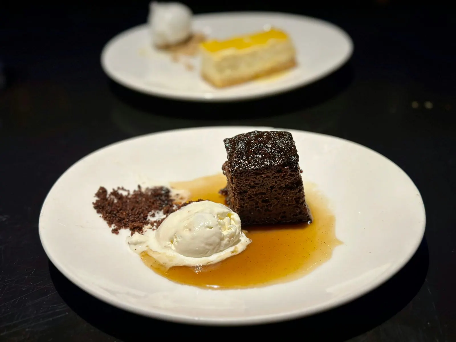 A square of dark sponge with ice cream, crumbs and caramel sauce, a slice of cheesecake behind it.