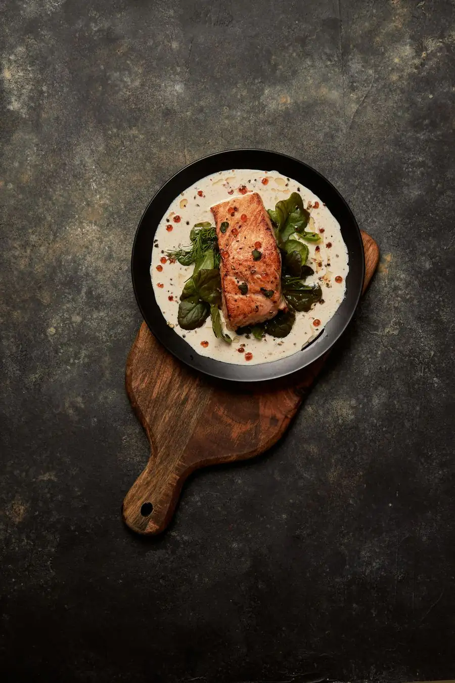A fillet of fish on a pale sauce with greens, on a dark plate and a wooden board, seen from above.