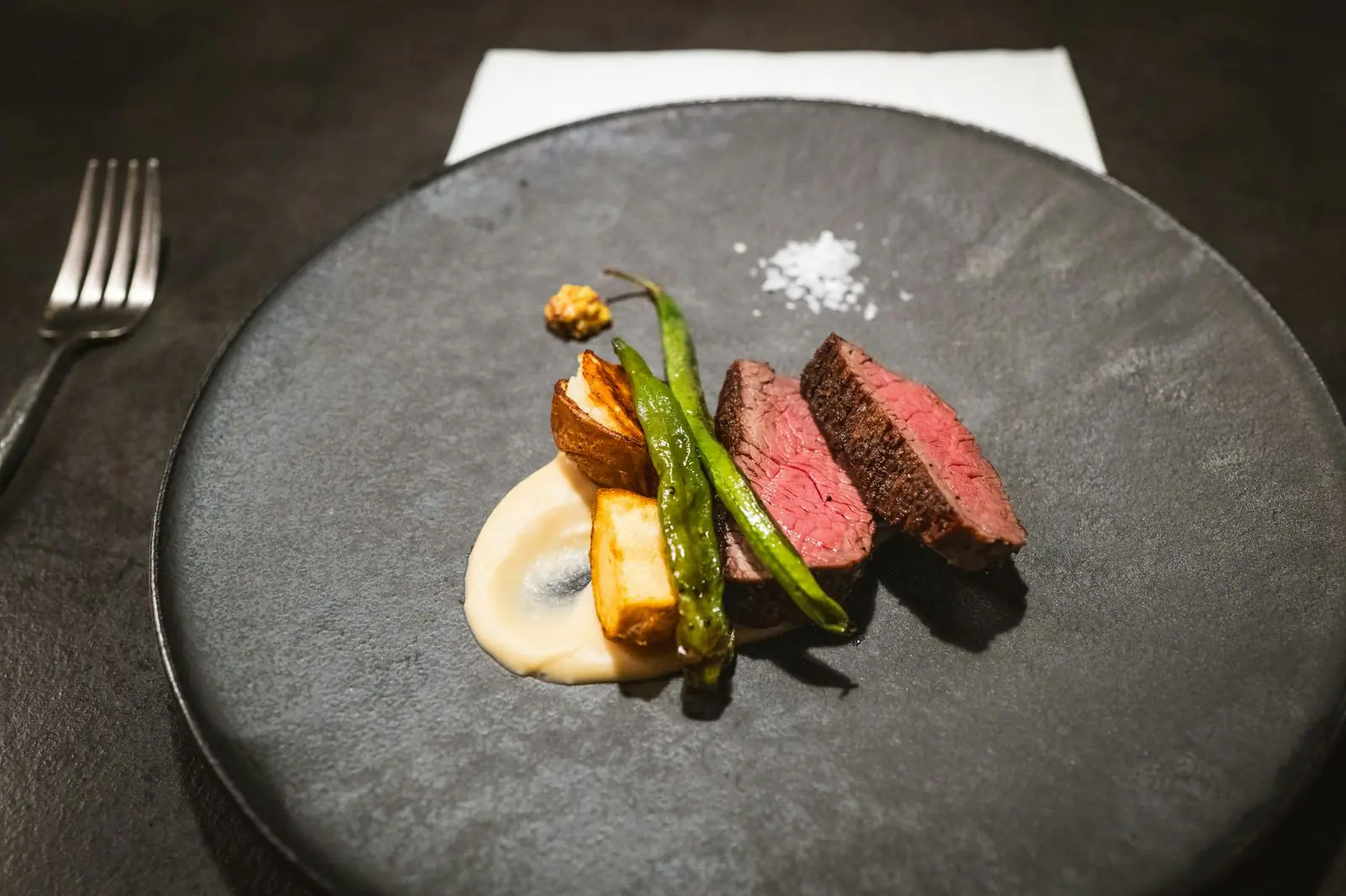 Slices of pink roast venison with green beans, roast potatoes and a pale puree on a dark stone plate.