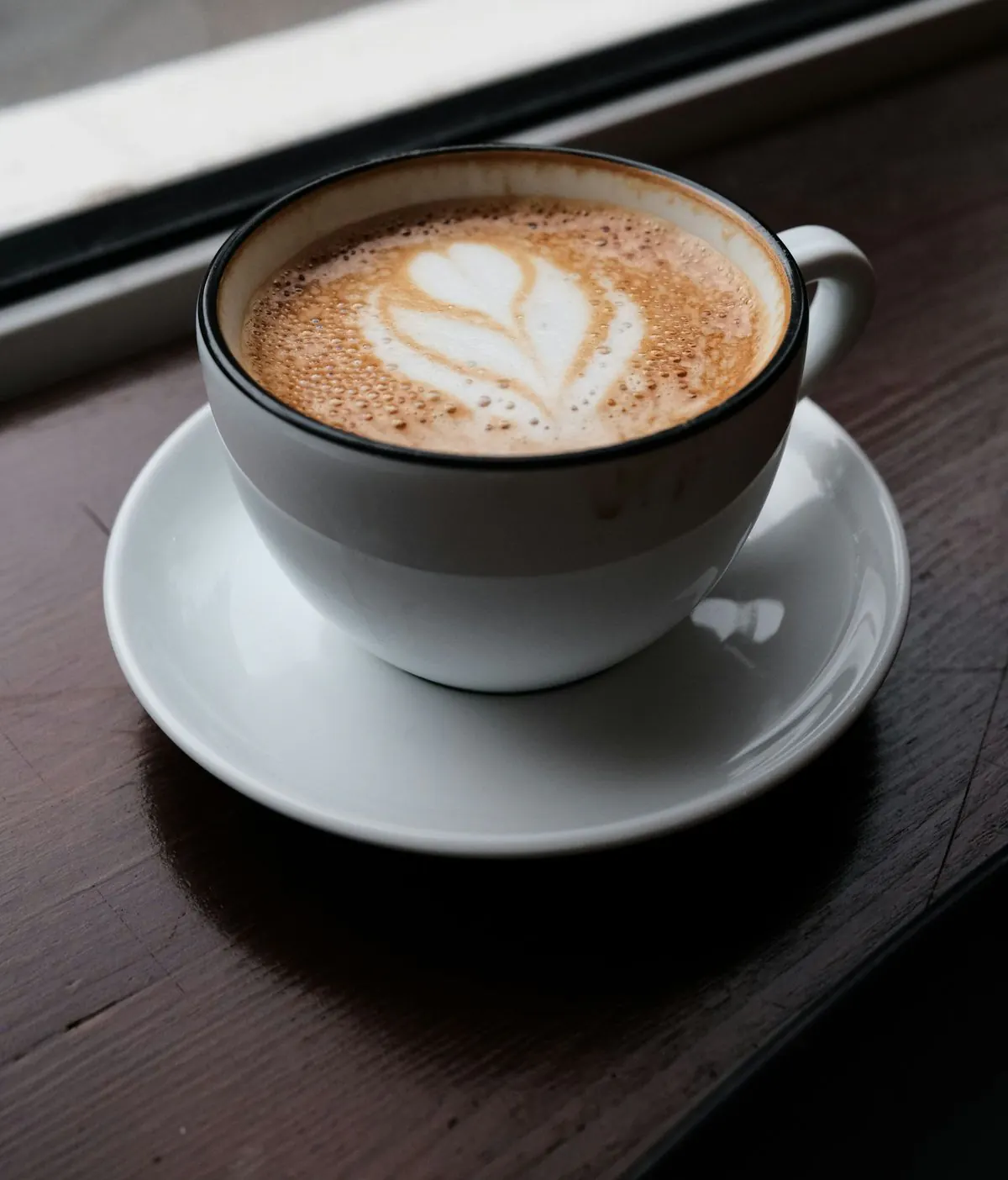 A flat white with a leaf drawn in the foam, on a saucer by the window.