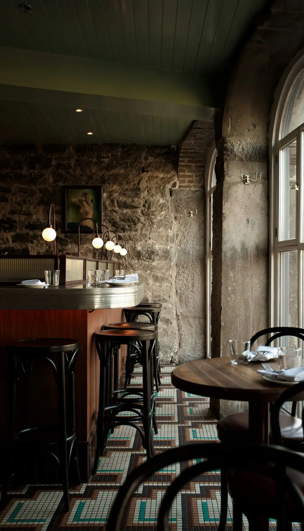 A corner of the dining room by the tall arched window, with a stone wall, bar stools, bentwood chairs and an old tiled floor.