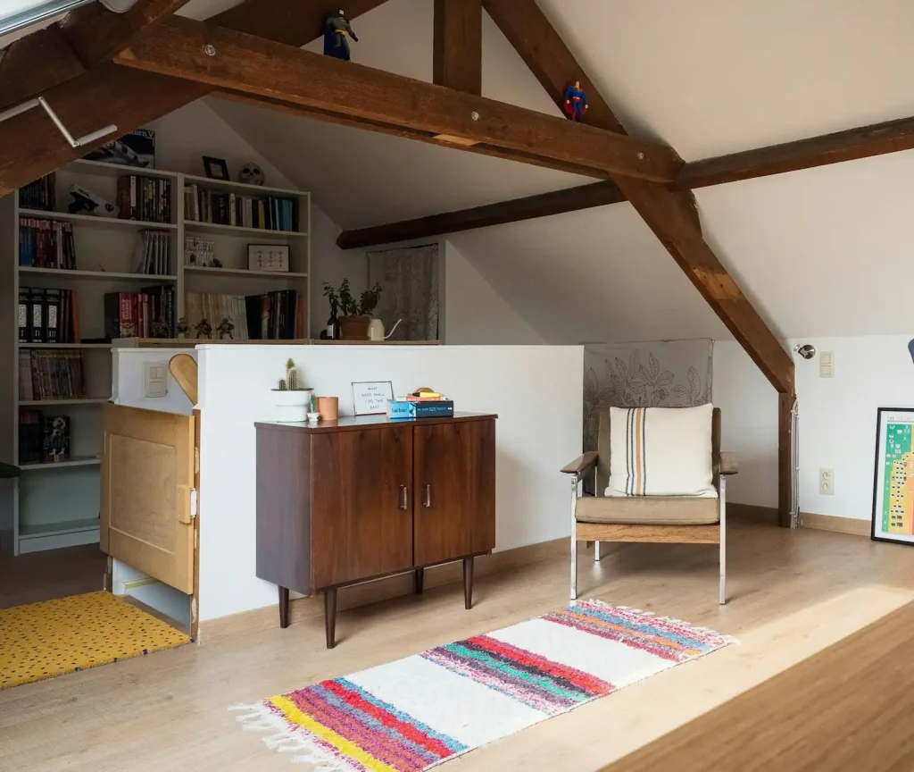 A bright attic room under a sloping ceiling with dark old collar beams, built in bookshelves, a wooden sideboard and a striped rug on a pale floor.