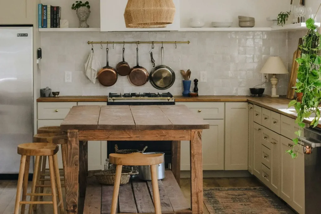 A kitchen with cream units, open shelves, copper pans on a rail and a rough oak island with two wooden stools in front of the cooker.
