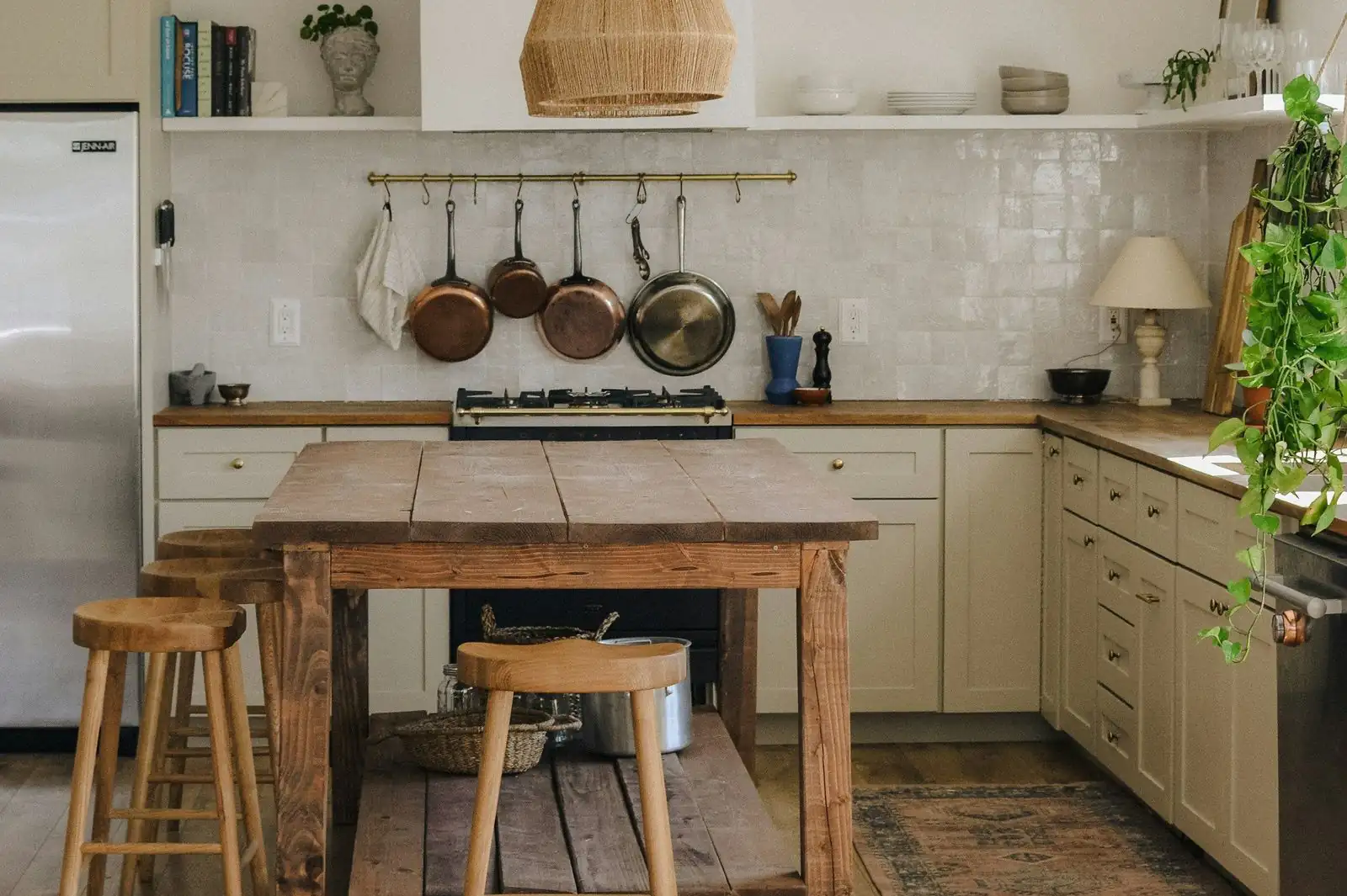 A kitchen with cream units, open shelves, copper pans on a rail and a rough oak island with two wooden stools in front of the cooker.