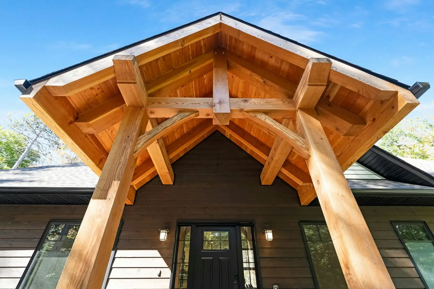 A new timber frame porch in front of a dark clad house: two heavy posts with curved braces carry an open gable of pale rafters.