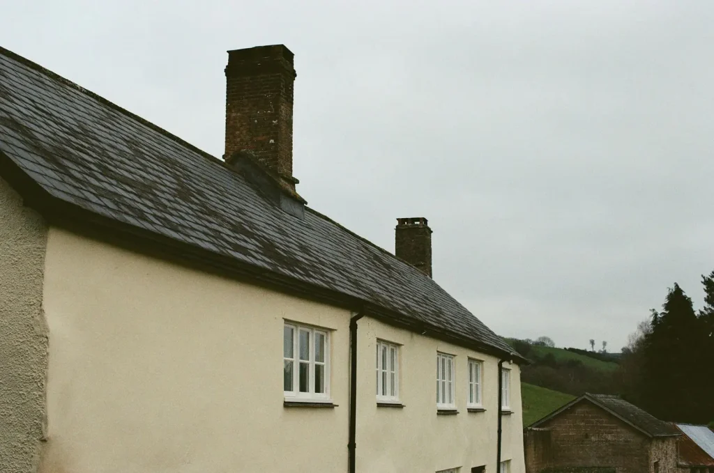 A long cream farmhouse with small white windows under a dark slate roof with two brick chimneys, green hills behind it.