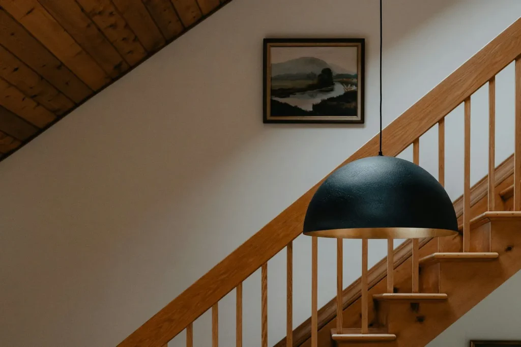A wooden staircase rising along a white wall, with a handrail on turned spindles, a black pendant lamp in front and a framed landscape on the wall.