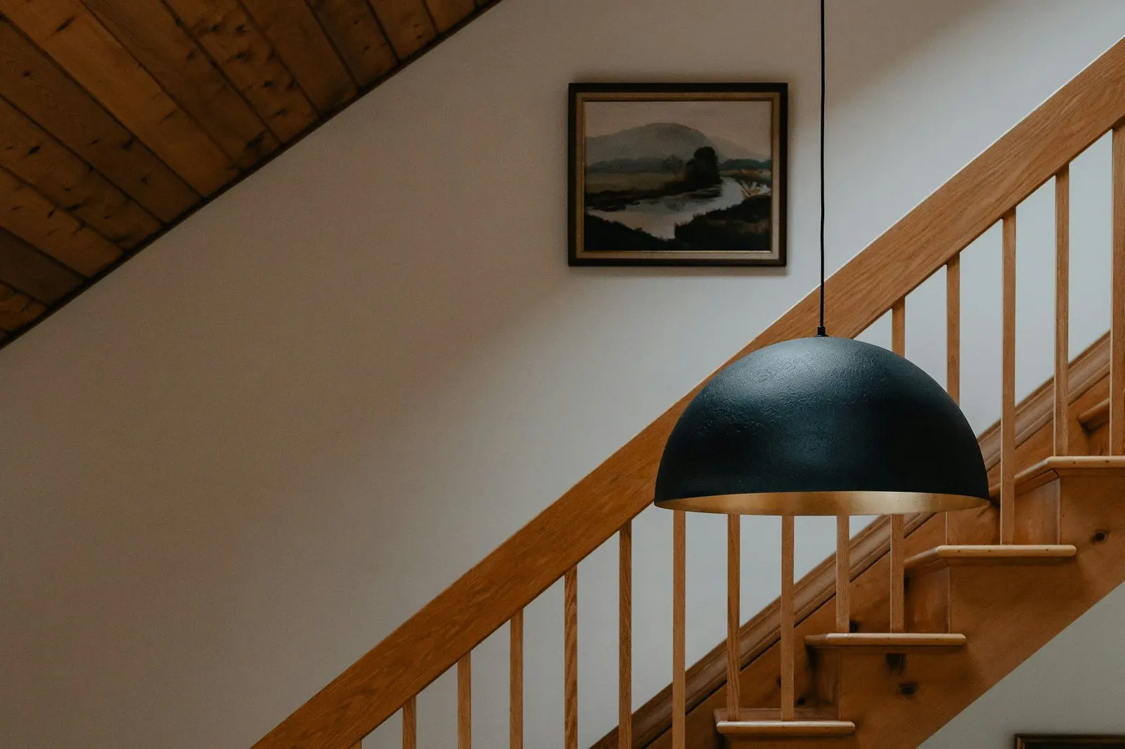 A wooden staircase rising along a white wall, with a handrail on turned spindles, a black pendant lamp in front and a framed landscape on the wall.